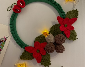 Könnte beinhalten: Ein grüner gehäkelter Kranz, der mit roten Weihnachtssternen, braunen Tannenzapfen und einem roten Glöckchen verziert ist. Der Kranz ist mit einer Lichterkette geschmückt, die einen festlichen Touch verleiht.