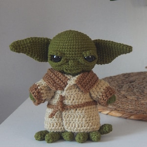 Pode incluir: Um brinquedo de pelúcia verde de crochê com orelhas grandes, olhos castanhos e um casaco de crochê bege. O brinquedo está em pé sobre uma superfície branca.