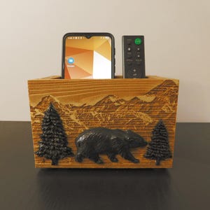 May include: Wooden organizer with a carved mountain scene and a black bear. It has two slots for holding a phone and a remote control.