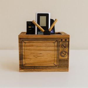 May include: Wooden TV-shaped remote control holder. The holder has a retro TV design with etched details. It contains several remote controls and a media player. The wood grain is visible, and the holder sits on a white surface.