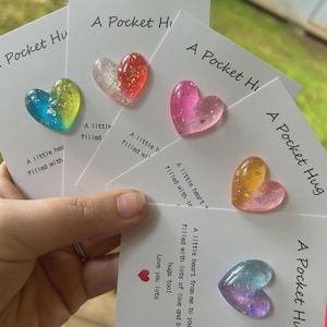 May include: Five white rectangular cards each featuring a small, glossy, heart-shaped resin charm with glitter accents. The cards are printed with the text A Pocket Hug and a message that reads A little heart filled with lots of love and big hugs too!