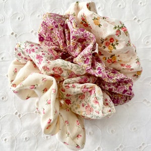 May include: A set of three fabric scrunchies in a floral pattern. The scrunchies are made of a soft, lightweight fabric and feature a variety of floral designs in shades of pink, cream, and purple.