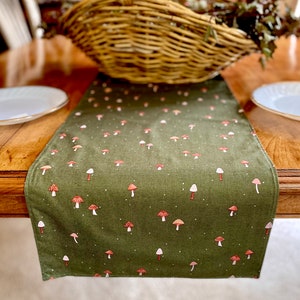 May include: A green table runner with a white background and a pattern of red and white mushrooms. The runner is on a wooden table with a wicker basket in the background.