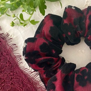 May include: A close-up of a burgundy scrunchie with black leopard print detailing. The scrunchie is made of a soft, textured fabric. A piece of burgundy lace is visible in the lower left corner. Green foliage is in the background.