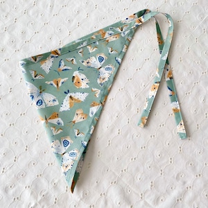 May include: A triangular bandana with a light green background and a repeating pattern of moths in white, brown, and blue. The bandana has matching ties for securing around the neck or head.