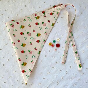 May include: A cream-colored triangular banner with a repeating pattern of strawberries in red, pink, and yellow. The banner has ties for hanging and is accompanied by several colorful buttons. The strawberries have smiling faces.