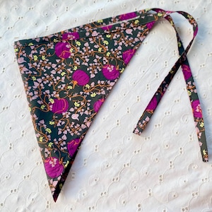 May include: A triangular bandana with a dark gray background, adorned with a pattern of purple pumpkins, orange vines, and small green and pink leaves. The bandana has long ties for securing it. The fabric is likely cotton or a similar material.