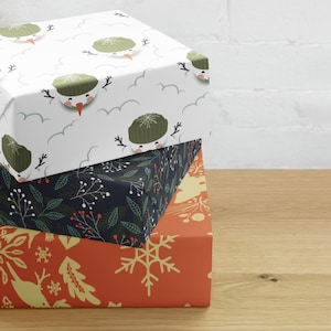 Peut inclure: Une pile de trois boîtes cadeaux rectangulaires enveloppées dans du papier à motifs. La boîte supérieure présente un fond blanc avec des bonhommes de neige portant des bonnets verts. La boîte centrale est bleu foncé avec des feuilles blanches et des baies rouges. La boîte inférieure est orange avec des flocons de neige dorés.