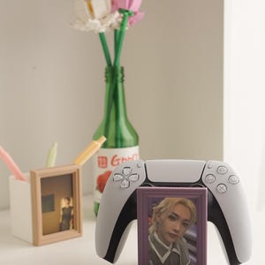 May include: A white and black PlayStation 5 controller with a purple framed photo in front. A green bottle holds Lego flowers. A white container holds pens and a framed photo.