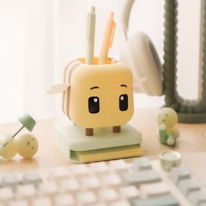 Könnte beinhalten: Ein gelber Schreibtisch-Organizer in Form einer Cartoonfigur mit einem lächelnden Gesicht, Flügeln und braunen Streifen. Zwei Stifte befinden sich in dem Organizer. Der Organizer steht auf einer hellgrünen Basis mit Haftnotizen. Eine Tastatur und Kopfhörer sind im Hintergrund.