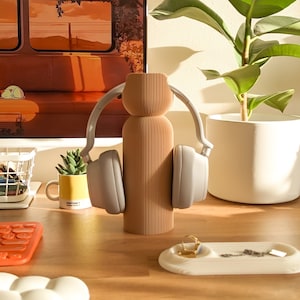 May include: A light brown, ribbed ceramic vase with a pair of grey over-ear headphones resting on it. The headphones are on a wooden desk with a small plant in a yellow mug, a keyboard, and a white tray with a gold ring and a silver chain.