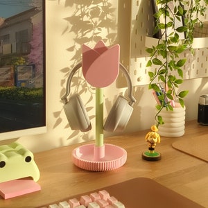 Puede incluir: Un soporte para auriculares en forma de tulip&aacute;n rosa con un tallo verde y unos auriculares grises. El soporte est&aacute; sobre un escritorio de madera con un teclado, un porta notas adhesivas en forma de rana verde y una peque&ntilde;a figura.