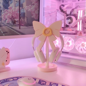 May include: A pink and yellow bow-shaped headphone stand with white headphones hanging from it. The stand is on a white desk with a white mouse and a pink and blue mousepad with a moon and stars design.