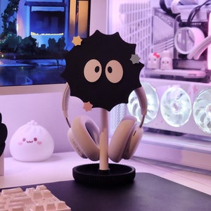 May include: A black headphone stand featuring a cartoon soot sprite design, supporting white headphones. The stand has a circular base and a central pole. The background includes a computer monitor, a computer case, and a keyboard.