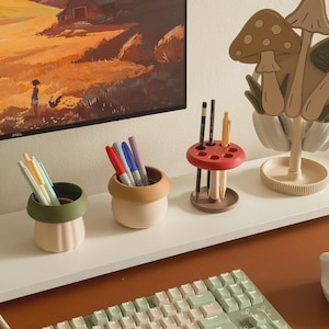 Op de afbeelding: Een verzameling bureau-organizers en decoraties. Inclusief een rode en bruine pennenhouder, tweekleurige beige en groene penhouders en een display met paddenstoelenthema. Een computermonitor toont een landschapsschilderij.