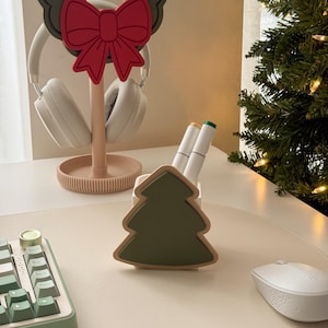 Puede incluir: Una configuración de escritorio festiva con un soporte para auriculares con forma de corona y un lazo rojo. También hay un portalápices con forma de árbol de Navidad verde, junto con un teclado, un ratón y un pequeño árbol de Navidad en el fondo.