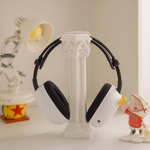 May include: White and black headphones resting on a white decorative column. The headphones have a modern design. A small desk lamp and a figurine are in the background.