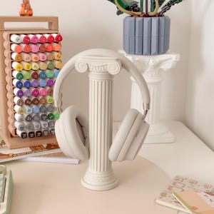 Puede incluir: Auriculares grises claros sobre un soporte de columna blanco decorativo. El soporte est&aacute; frente a un escritorio con una alfombrilla rosa claro. Tambi&eacute;n se ve un estuche de rotuladores de colores y una planta de Lego.