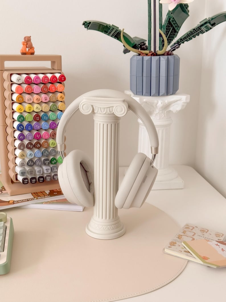 Puede incluir: Auriculares grises claros sobre un soporte de columna blanco decorativo. El soporte est&aacute; frente a un escritorio con una alfombrilla rosa claro. Tambi&eacute;n se ve un estuche de rotuladores de colores y una planta de Lego.