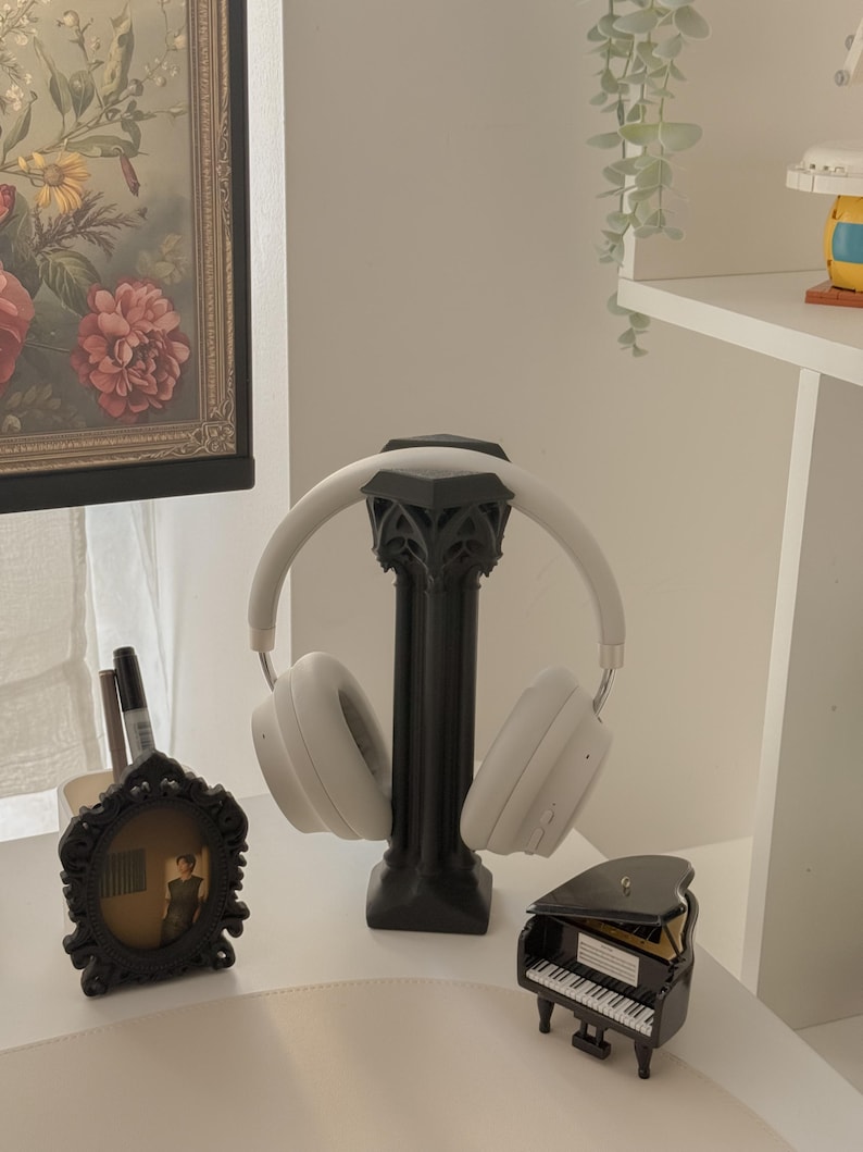 May include: White wireless headphones on a black decorative stand. A small black framed picture and a miniature black piano are also on display. The background includes a floral painting and a white shelf.