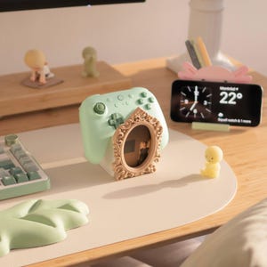 May include: A light green gaming controller stand featuring an ornate, oval-shaped beige frame on the front. The stand is designed to hold a controller and includes a small, central display area within the decorative frame for a photograph.