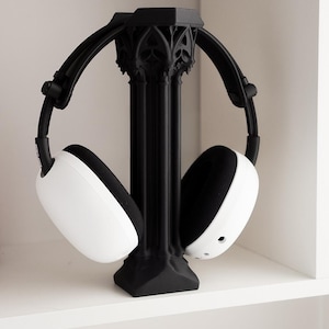 May include: Black headphone stand with a gothic architectural design, holding white and black headphones. The stand has a central column and arched arms to cradle the headphones. The headphones have white earcups and black padding.