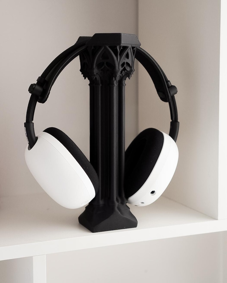 May include: Black headphone stand with a gothic architectural design, holding white and black headphones. The stand has a central column and arched arms to cradle the headphones. The headphones have white earcups and black padding.