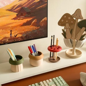 Pu&ograve; includere: Una collezione di organizzatori e accessori da scrivania. Include un supporto per cuffie a forma di fungo, portapenne di vari colori e una tastiera. I portapenne hanno la forma di piccoli vasi, con penne e pennarelli all'interno.
