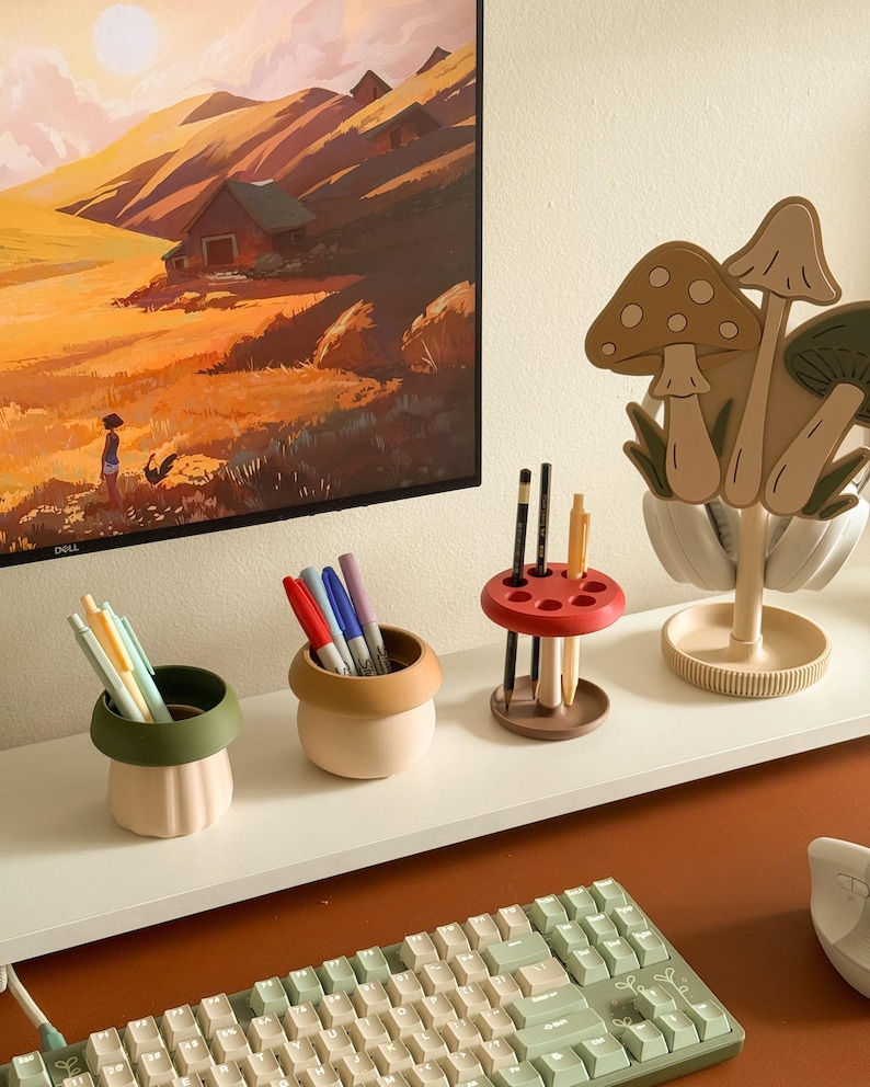 Pu&ograve; includere: Una collezione di organizzatori e accessori da scrivania. Include un supporto per cuffie a forma di fungo, portapenne di vari colori e una tastiera. I portapenne hanno la forma di piccoli vasi, con penne e pennarelli all'interno.