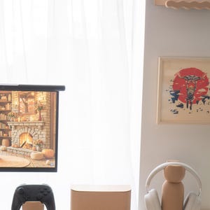 May include: A modern home entertainment setup featuring a black game controller, a brown speaker, and a headphone stand. The stand holds white headphones. A screen displays a fireplace scene. The items are on a white surface.