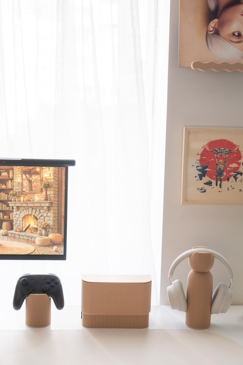 May include: A modern home entertainment setup featuring a black game controller, a brown speaker, and a headphone stand. The stand holds white headphones. A screen displays a fireplace scene. The items are on a white surface.