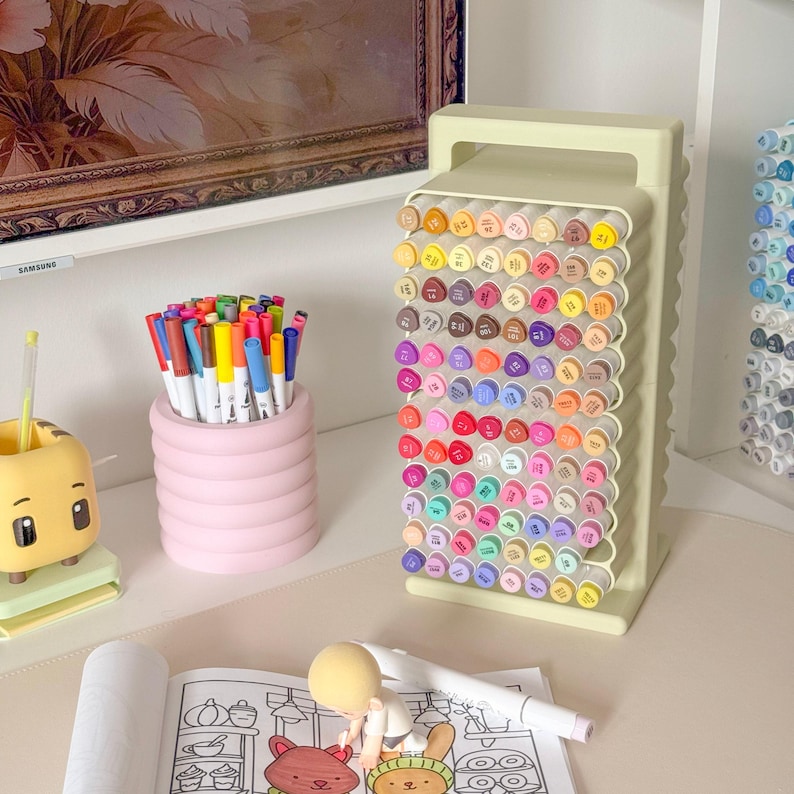 May include: A collection of art supplies, including a light green marker holder filled with colorful markers. A pink cylindrical pen holder is filled with various colored pens. A coloring book and a small figurine are also present.