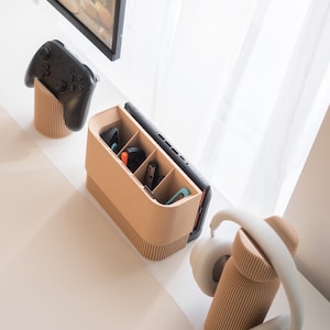 May include: A beige, ribbed-textured organizer holds a black game controller, a smartphone, and various game accessories. A pair of white headphones rests on a matching ribbed stand. The items are arranged on a white surface.