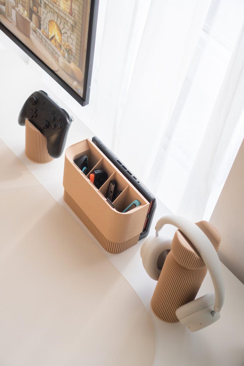 May include: A beige, ribbed-textured organizer holds a black game controller, a smartphone, and various game accessories. A pair of white headphones rests on a matching ribbed stand. The items are arranged on a white surface.