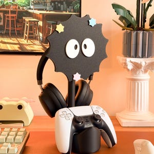 May include: A black headphone stand in the shape of a soot sprite, with black headphones and a white and black game controller. The stand features white eyes and star accents. A keyboard and frog-shaped desk tidy are also visible.