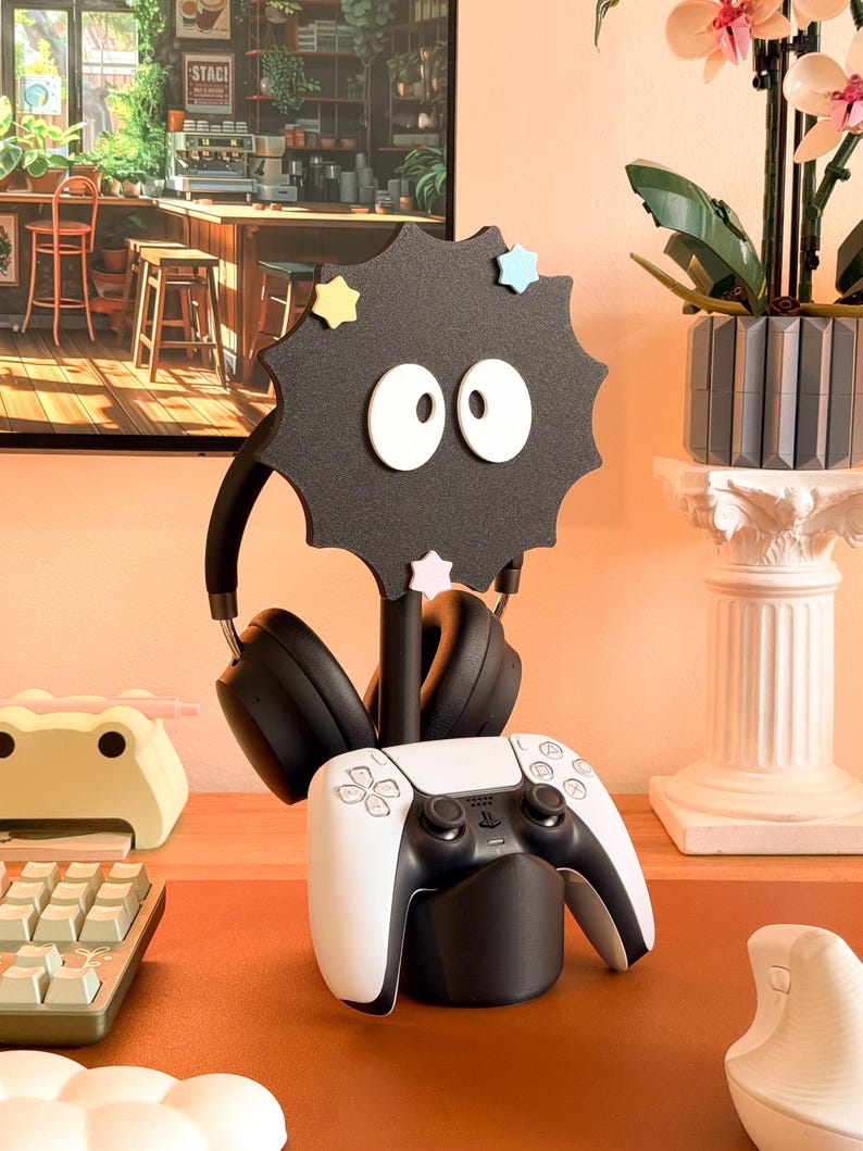 May include: A black headphone stand in the shape of a soot sprite, with black headphones and a white and black game controller. The stand features white eyes and star accents. A keyboard and frog-shaped desk tidy are also visible.