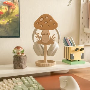 Pu&ograve; includere: Un supporto per cuffie a forma di fungo beige con base rotonda, che tiene cuffie bianche. Sono visibili anche un piccolo oggetto decorativo a tema fungo e un portapenne a tema ape. Una tastiera e un mouse sono in primo piano.