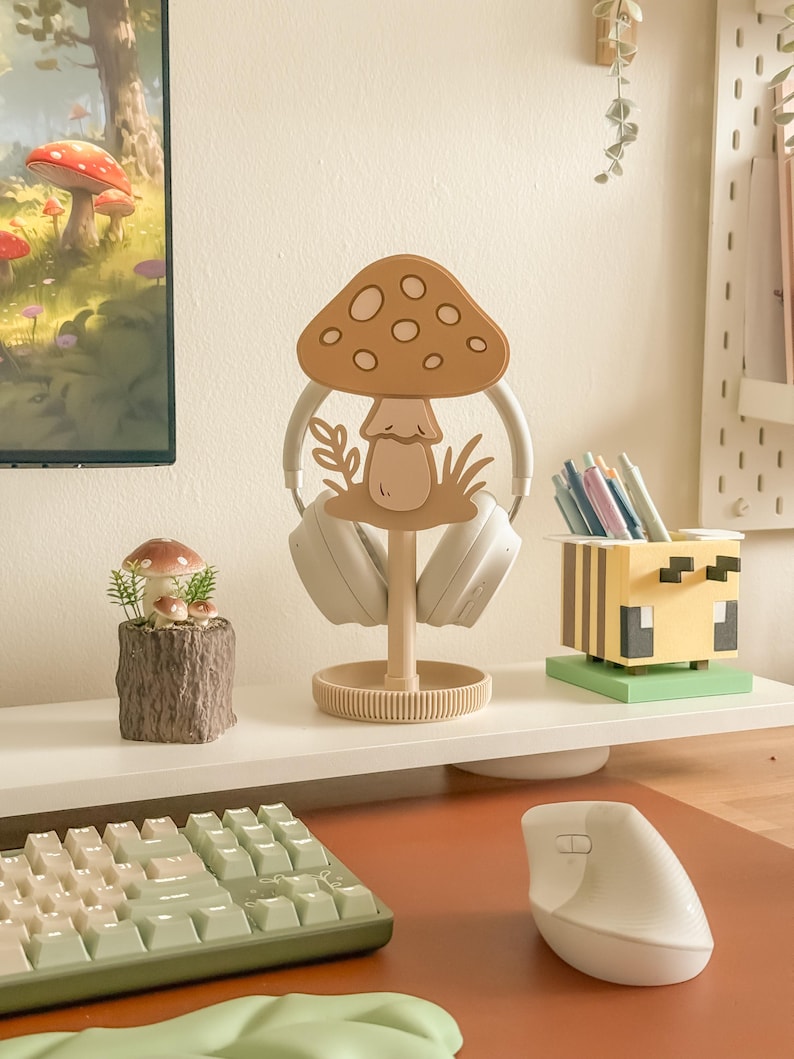 Pu&ograve; includere: Un supporto per cuffie a forma di fungo beige con base rotonda, che tiene cuffie bianche. Sono visibili anche un piccolo oggetto decorativo a tema fungo e un portapenne a tema ape. Una tastiera e un mouse sono in primo piano.