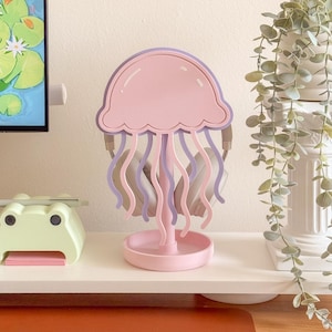 May include: A pink and purple jellyfish-shaped headphone stand. The stand has a round base and a central stem supporting the jellyfish design. Headphones are hanging from the stand. The stand is on a white shelf.