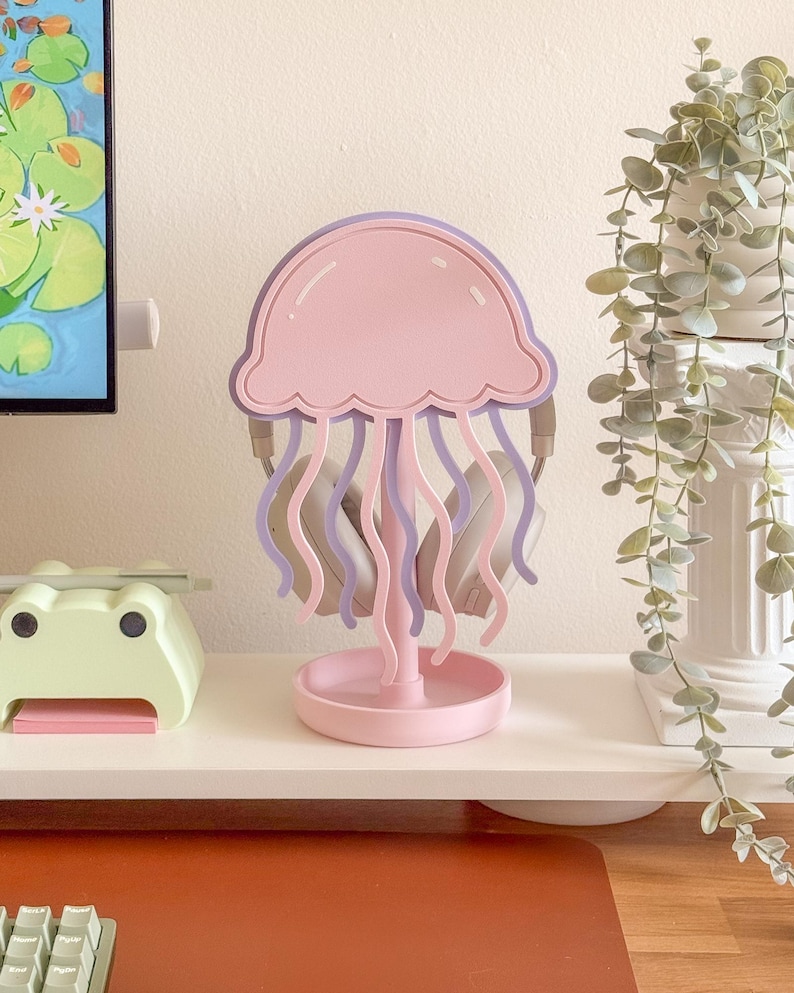 May include: A pink and purple jellyfish-shaped headphone stand. The stand has a round base and a central stem supporting the jellyfish design. Headphones are hanging from the stand. The stand is on a white shelf.