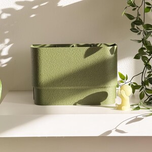 May include: A textured, olive green rectangular storage container. The container has a slightly rounded shape and is sitting on a white shelf. A small yellow candle and greenery are visible in the background.