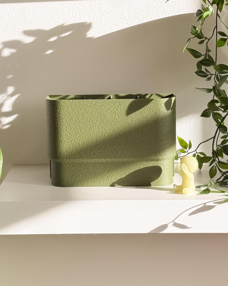 May include: A textured, olive green rectangular storage container. The container has a slightly rounded shape and is sitting on a white shelf. A small yellow candle and greenery are visible in the background.