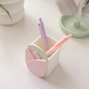 May include: A white desk organizer with a tulip design in pastel pink and green, holding a purple and a pink pen. The organizer is square-shaped and sits on a white surface, with a plant and headphones in the background.