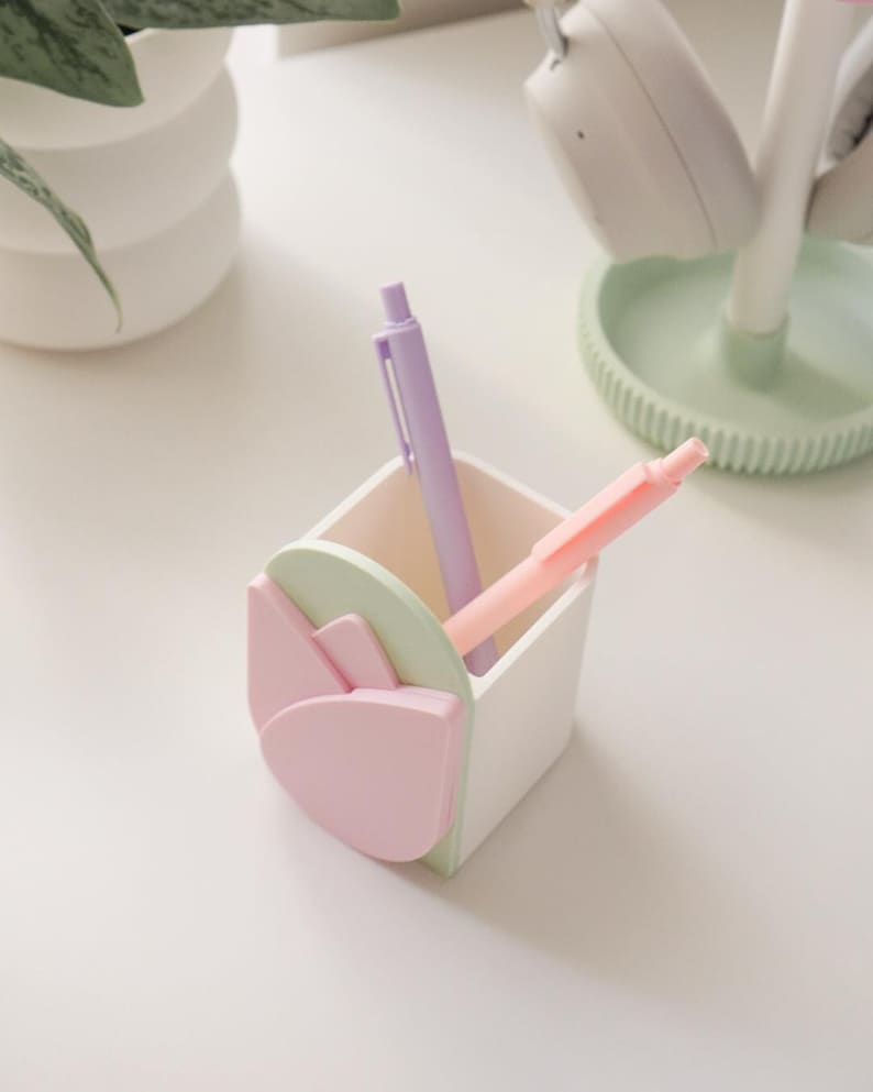 May include: A white desk organizer with a tulip design in pastel pink and green, holding a purple and a pink pen. The organizer is square-shaped and sits on a white surface, with a plant and headphones in the background.