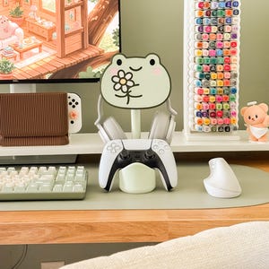 May include: A gaming setup featuring a white and black game controller resting on a light green stand. A frog-shaped headphone holder with a flower design is in the center. A keyboard, mouse, and marker holder are also visible.