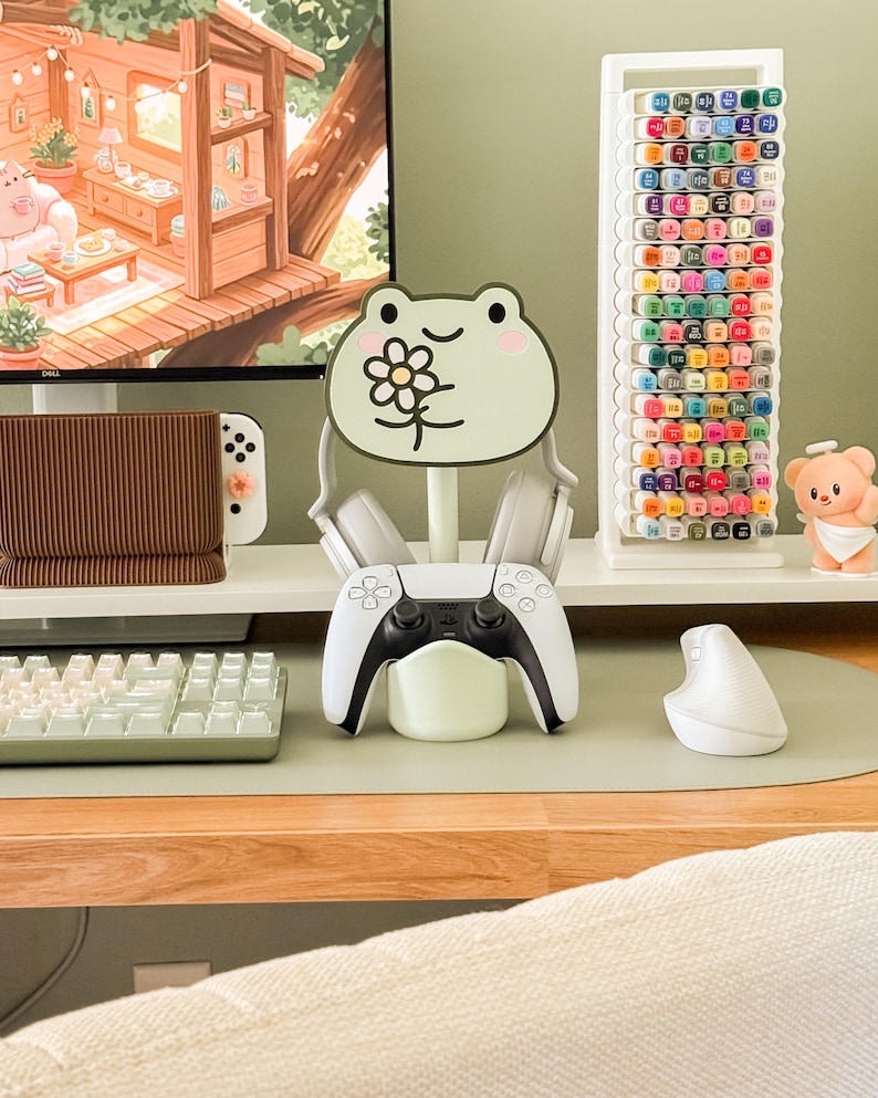 May include: A gaming setup featuring a white and black game controller resting on a light green stand. A frog-shaped headphone holder with a flower design is in the center. A keyboard, mouse, and marker holder are also visible.