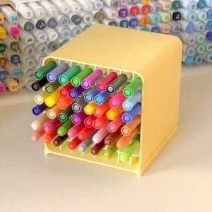 May include: A yellow square plastic desk organizer filled with a variety of colorful markers. The grid-style holder keeps each marker upright and separated. Additional organizers in white, tan, and light blue are visible in the background.