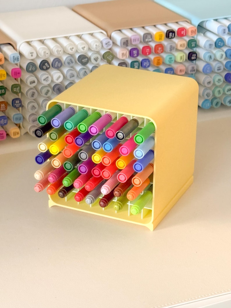 May include: A yellow square plastic desk organizer filled with a variety of colorful markers. The grid-style holder keeps each marker upright and separated. Additional organizers in white, tan, and light blue are visible in the background.
