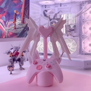 May include: A pink and white controller stand with a heart-shaped design and wings. The stand is holding a pink Xbox controller.