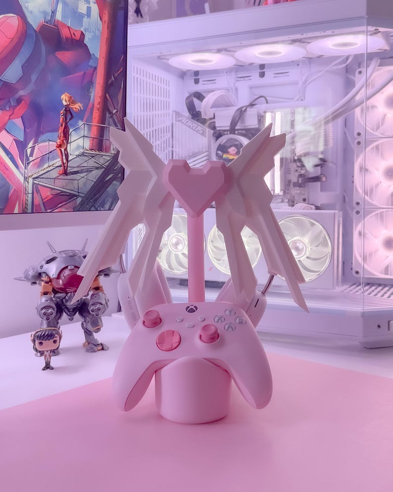 May include: A pink and white controller stand with a heart-shaped design and wings. The stand is holding a pink Xbox controller.